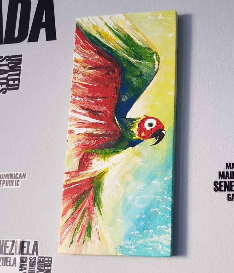 Akrylowe malowidło na płótnie przedstawiające papugę w locie, z przewagą czerwieni i zieleni, zawieszone na białej ścianie obok naklejek z nazwami państw.