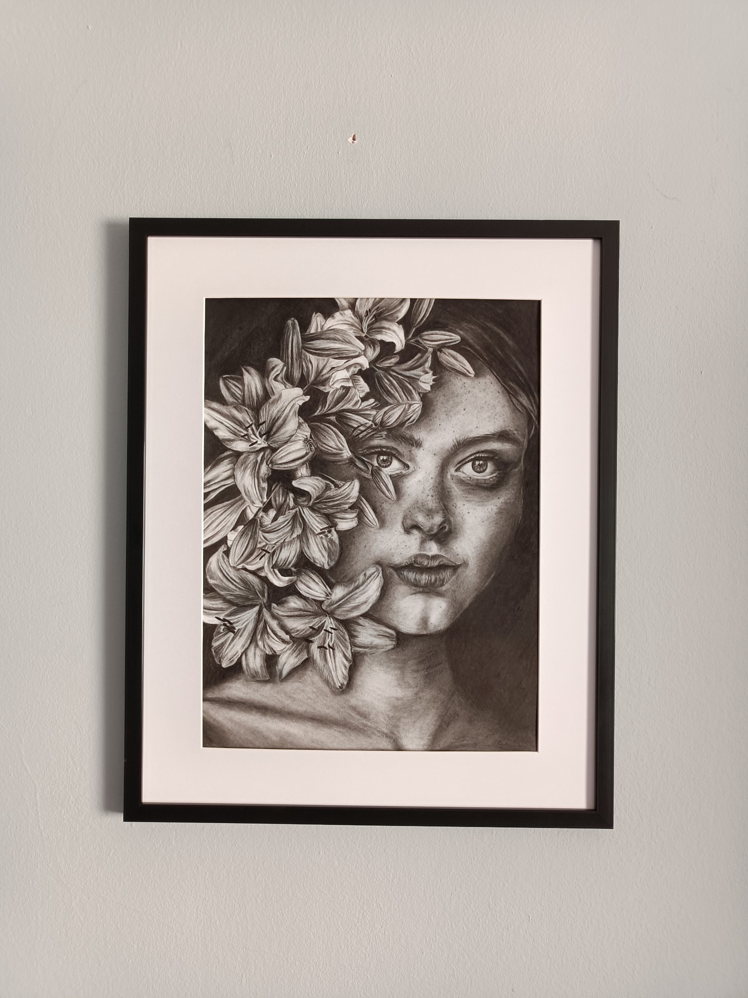 Oprawiony w ramę portret kobiety z kwiatami lilii w czerni i bieli, wiszący na jasnoszarej ścianie.