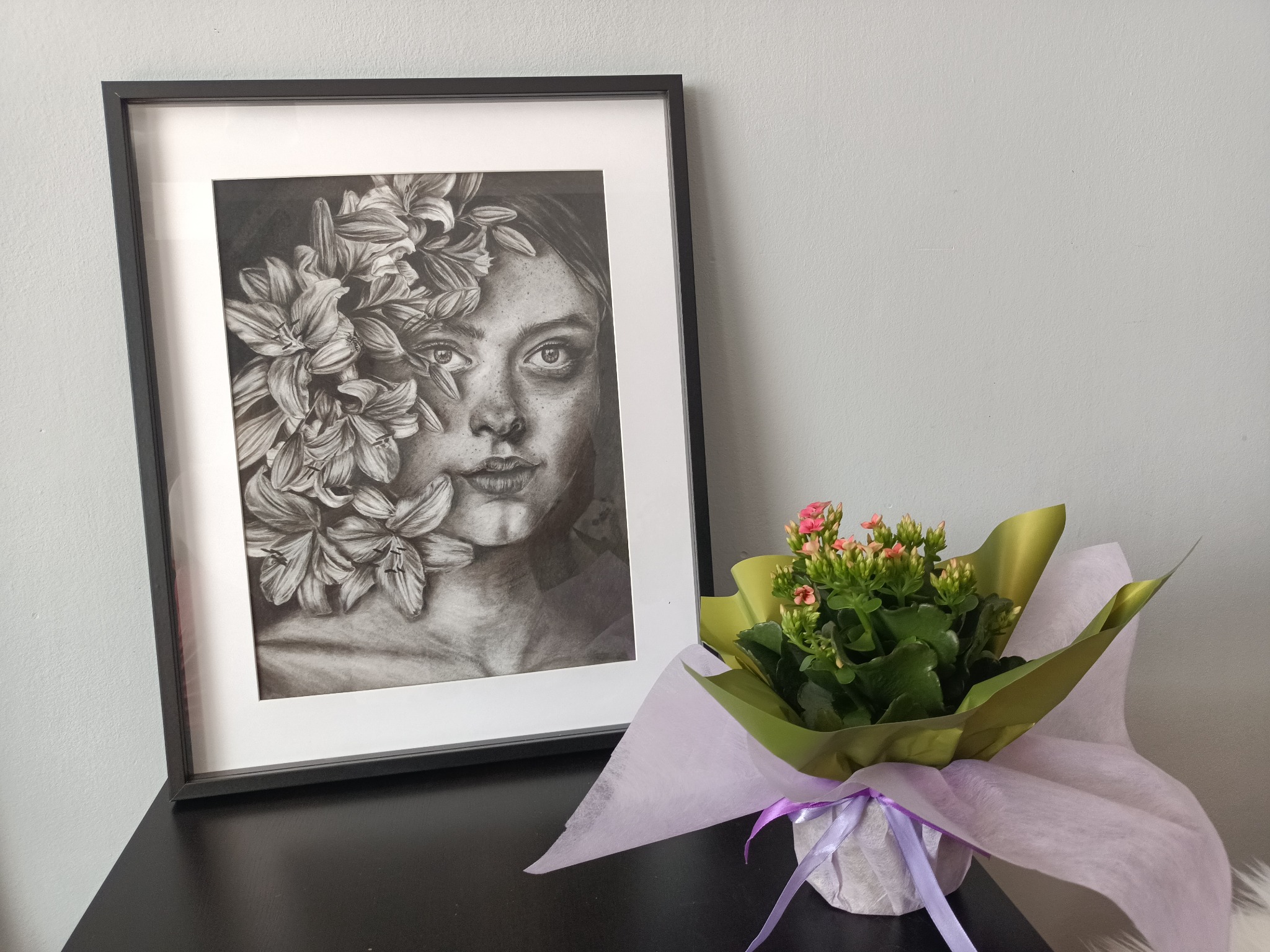 Oprawiony w ramkę portret kobiety z kwiatami w czerni i bieli, obok doniczka z kwitnącą kalonchoe w ozdobnym opakowaniu na czarnym blacie