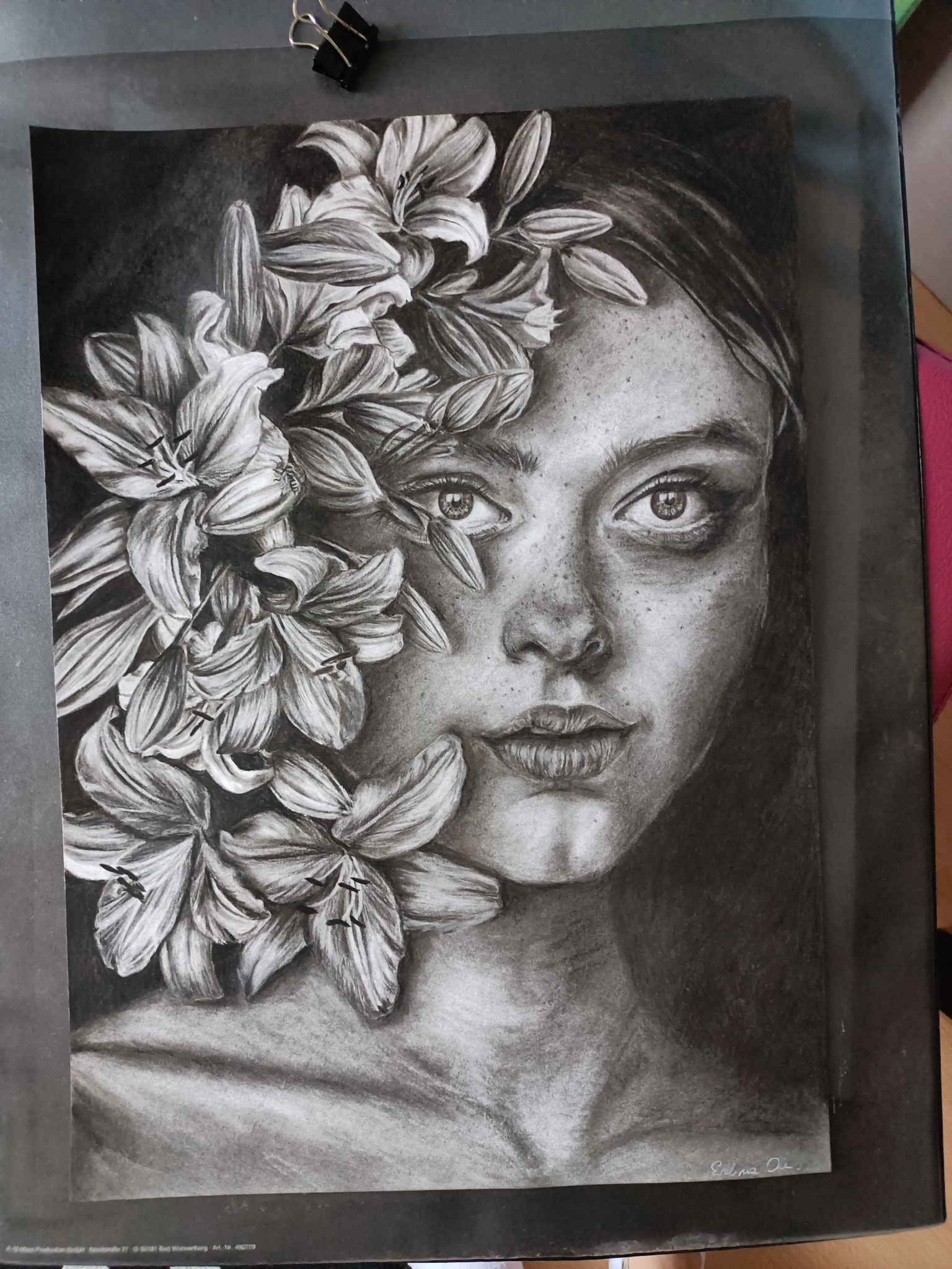 Czarno-biały portret kobiety z liliami, wykonany techniką rysunku ołówkiem, z widocznymi detalami twarzy i kwiatów, umieszczony na czarnym papierze z metalowym klipsem w rogu.