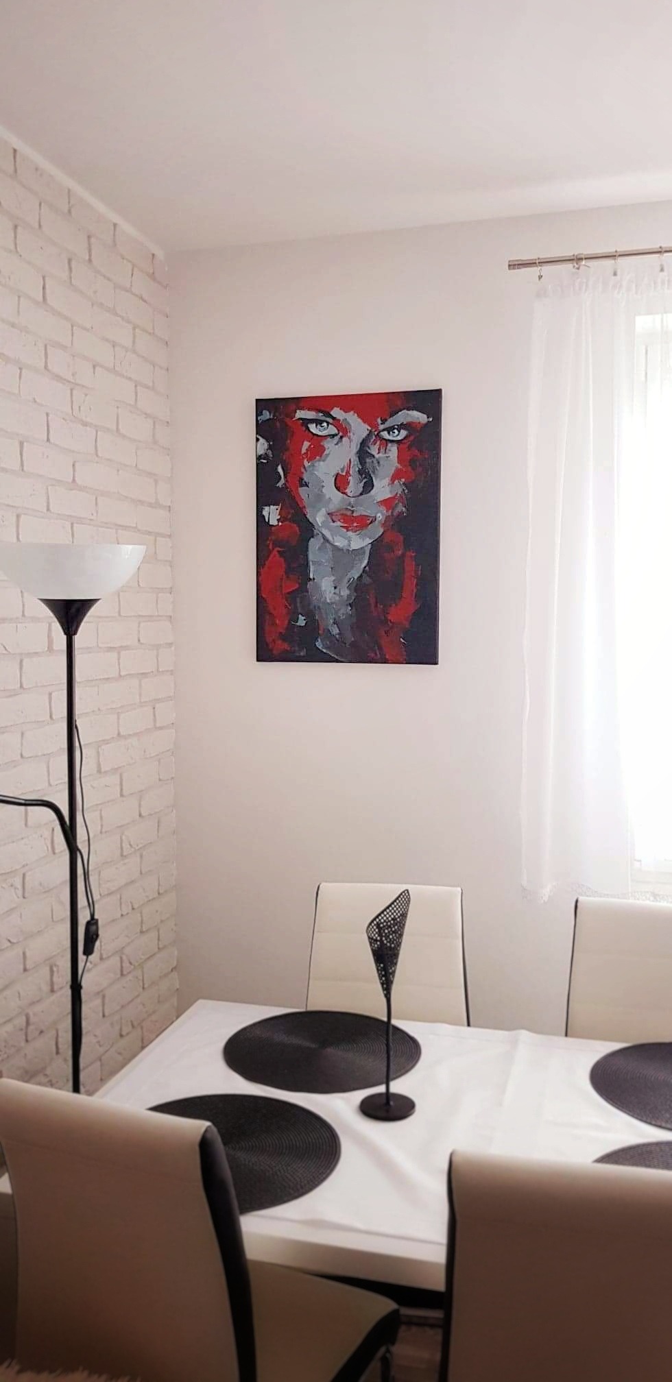 Abstrakcyjny portret kobiety w odcieniach czerwieni i szarości, wiszący na białej ścianie nad stołem w jadalni z białym obrusem i czarnymi podkładkami.