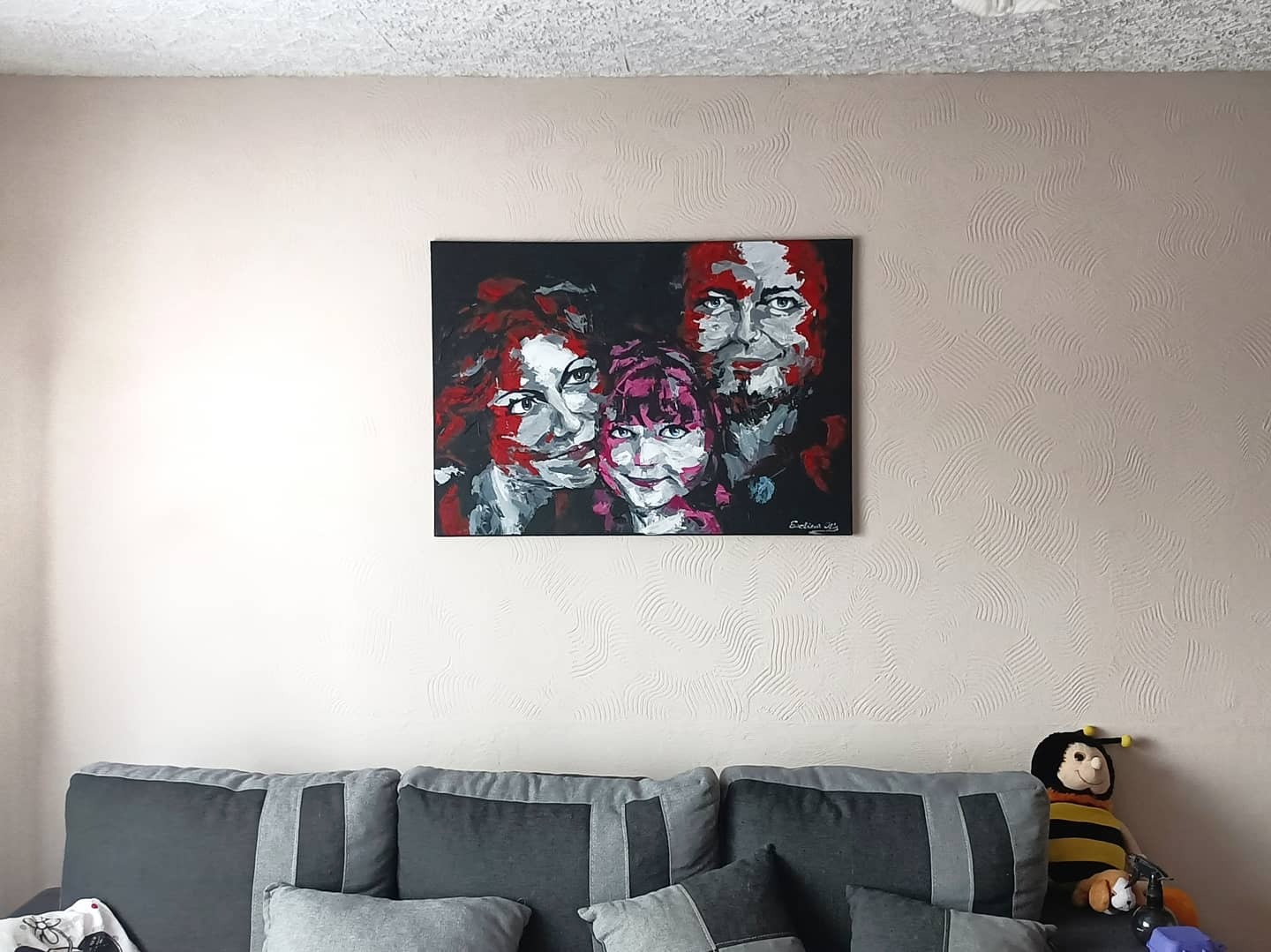 Abstrakcyjny portret rodzinny z użyciem czerwieni, bieli i czerni, wiszący nad szarą kanapą z poduszkami i pluszową pszczołą.
