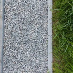 Ścieżka z drobnego, szarego żwiru, obramowana betonowym krawężnikiem i zieloną trawą.