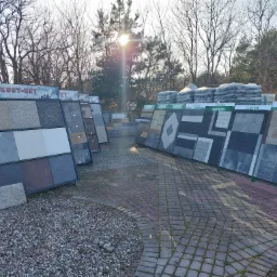 Ekspozycja różnorodnych wzorów i kolorów nawierzchni brukowych, prezentowana na stojakach firmowych Kost-Bet i Ginter, na tle placu wyłożonego kostką brukową i drzew bezlistnych w słoneczny dzień.