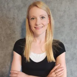 Portret uśmiechniętej blondynki z rękami skrzyżowanymi na tle szarej ściany.