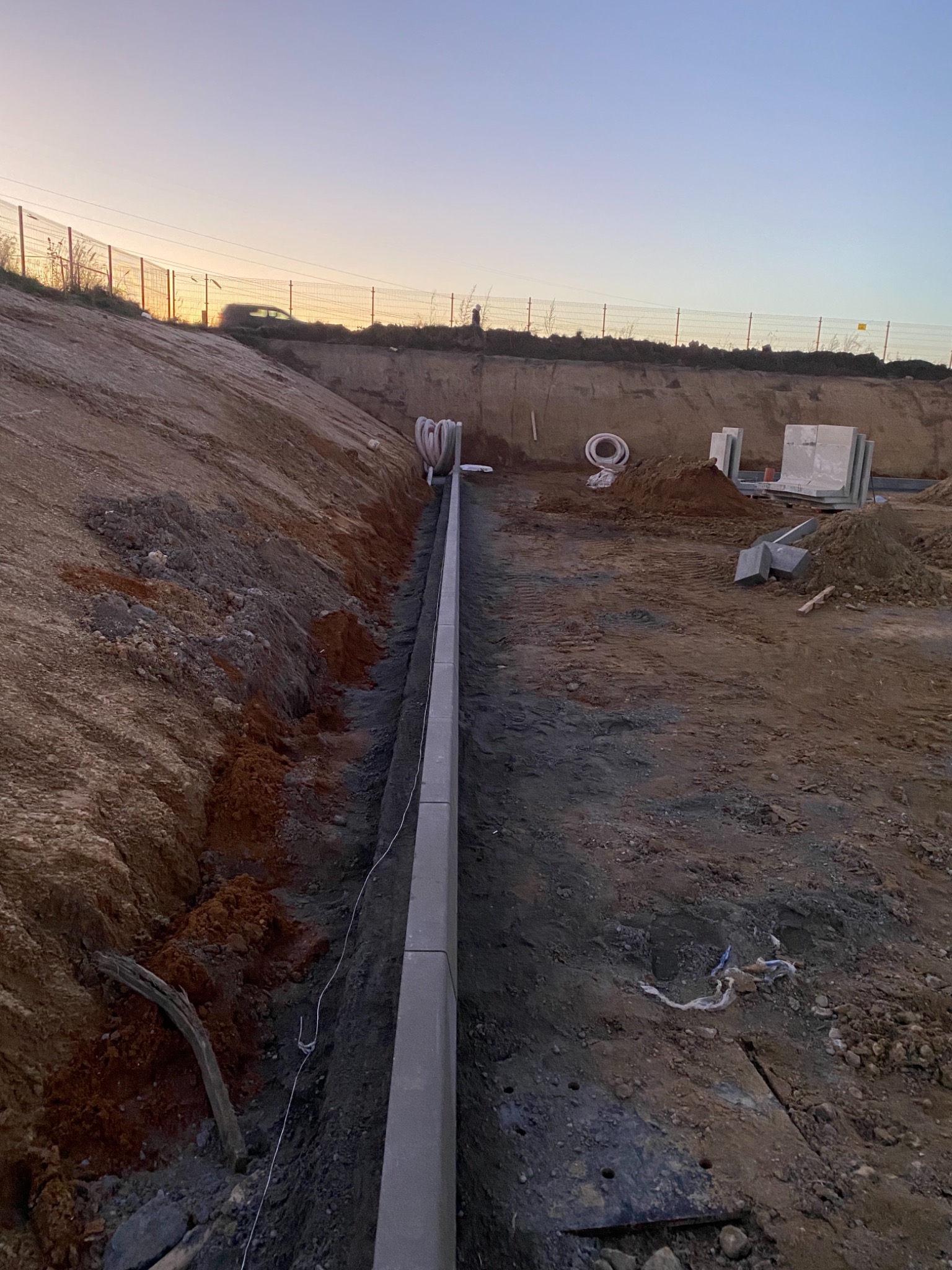 Świeżo ułożony obrzeżnik betonowy oddzielający wykop od terenu budowy, z widocznymi rurami i stertą płyt betonowych w tle, sfotografowane o zmierzchu.