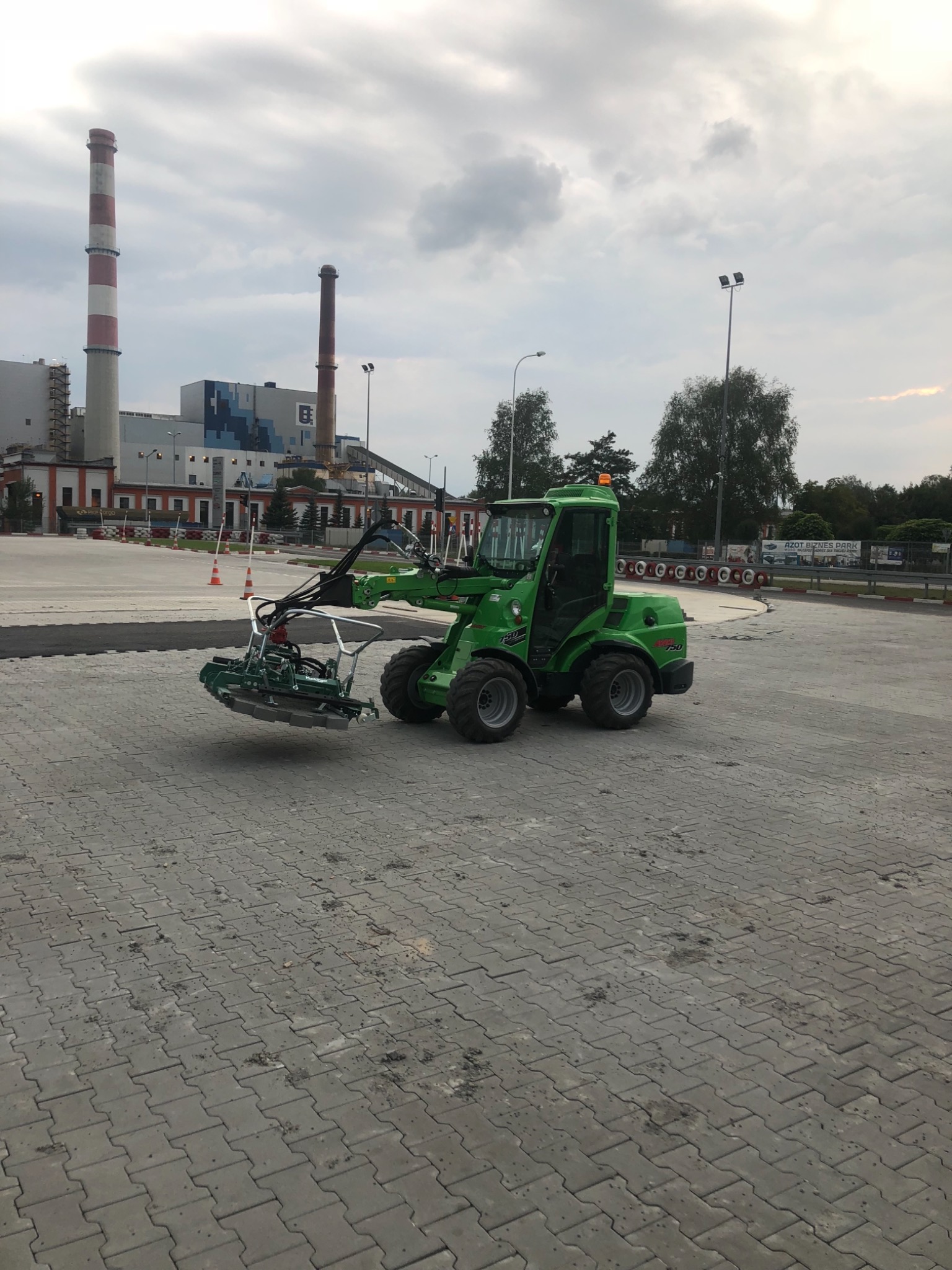 Zielona maszyna do układania kostki brukowej na placu, w tle kominy przemysłowe i pochmurne niebo.