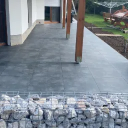 Zadaszony taras z szarych płyt, obudowany gabionem wypełnionym kamieniami, widok na zadbany ogr&oacute;d z suszarką na pranie i drewnianym stołem piknikowym.