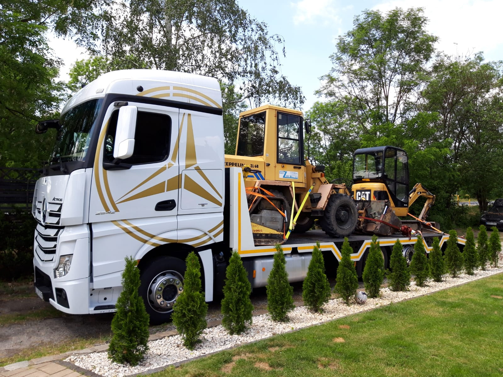 Biała laweta z ozdobnym złotym wzorem przewozi żółtą ładowarkę Zeppelin ZL 6B i mini koparkę CAT, zabezpieczone pasami transportowymi, na tle zieleni drzew i trawnika z ozdobnymi tujami.