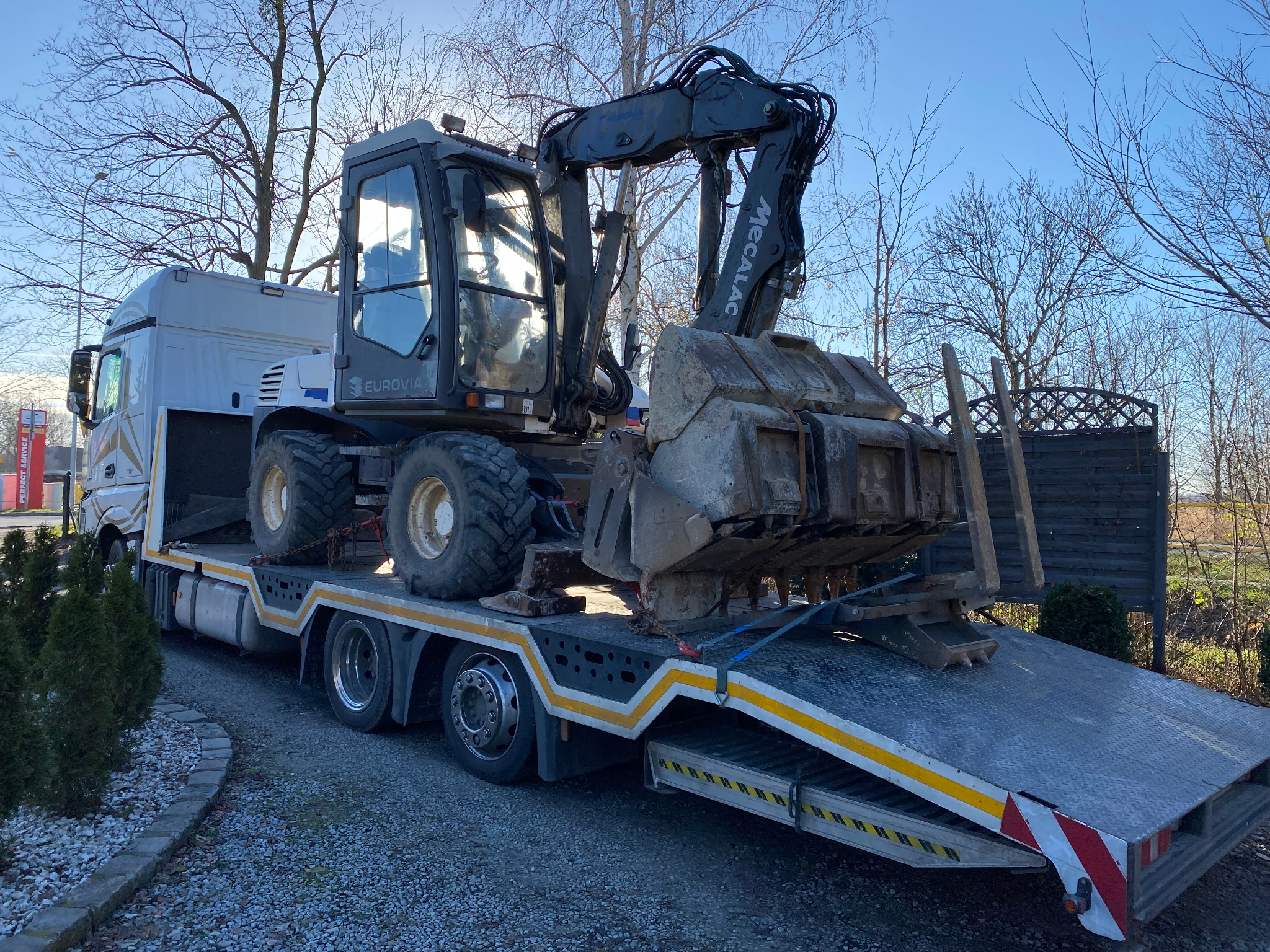Kołowa koparka Mecalac załadowana na lawetę ciężarówki Eurovia, zabezpieczona pasami transportowymi, na tle drzew i błękitnego nieba.