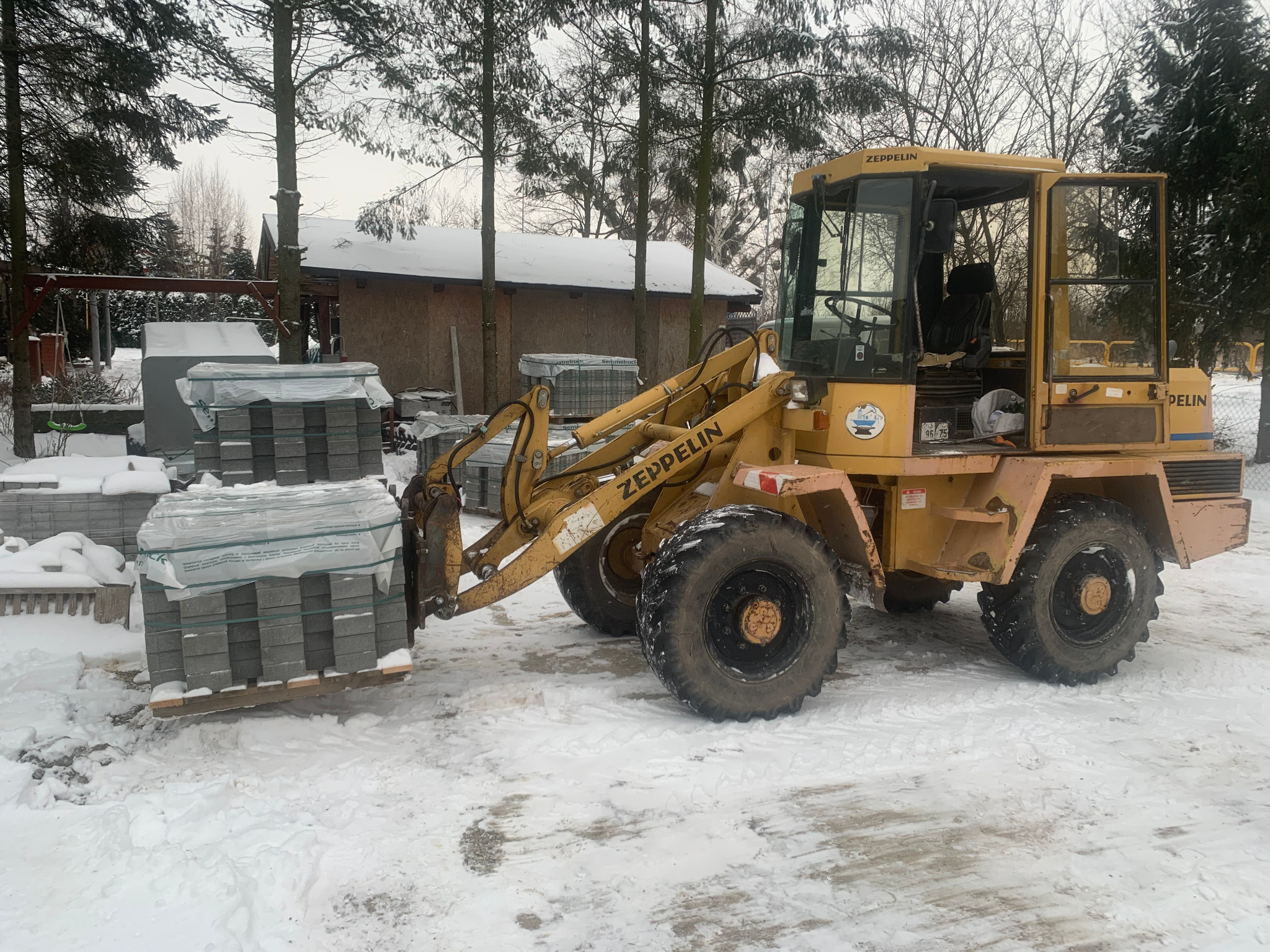 Żółta ładowarka Zeppelin przewozi paletę z kostką brukową w zimowej scenerii, na śniegu, z budynkiem w tle.