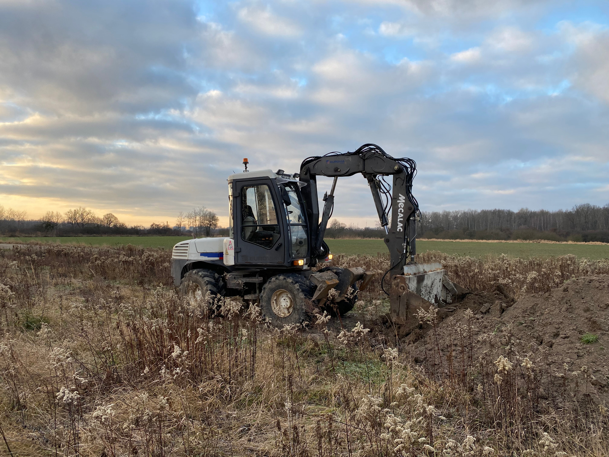 Koparka Mecalac podczas pracy w polu, wykopany dół, jesienna trawa, pochmurne niebo.
