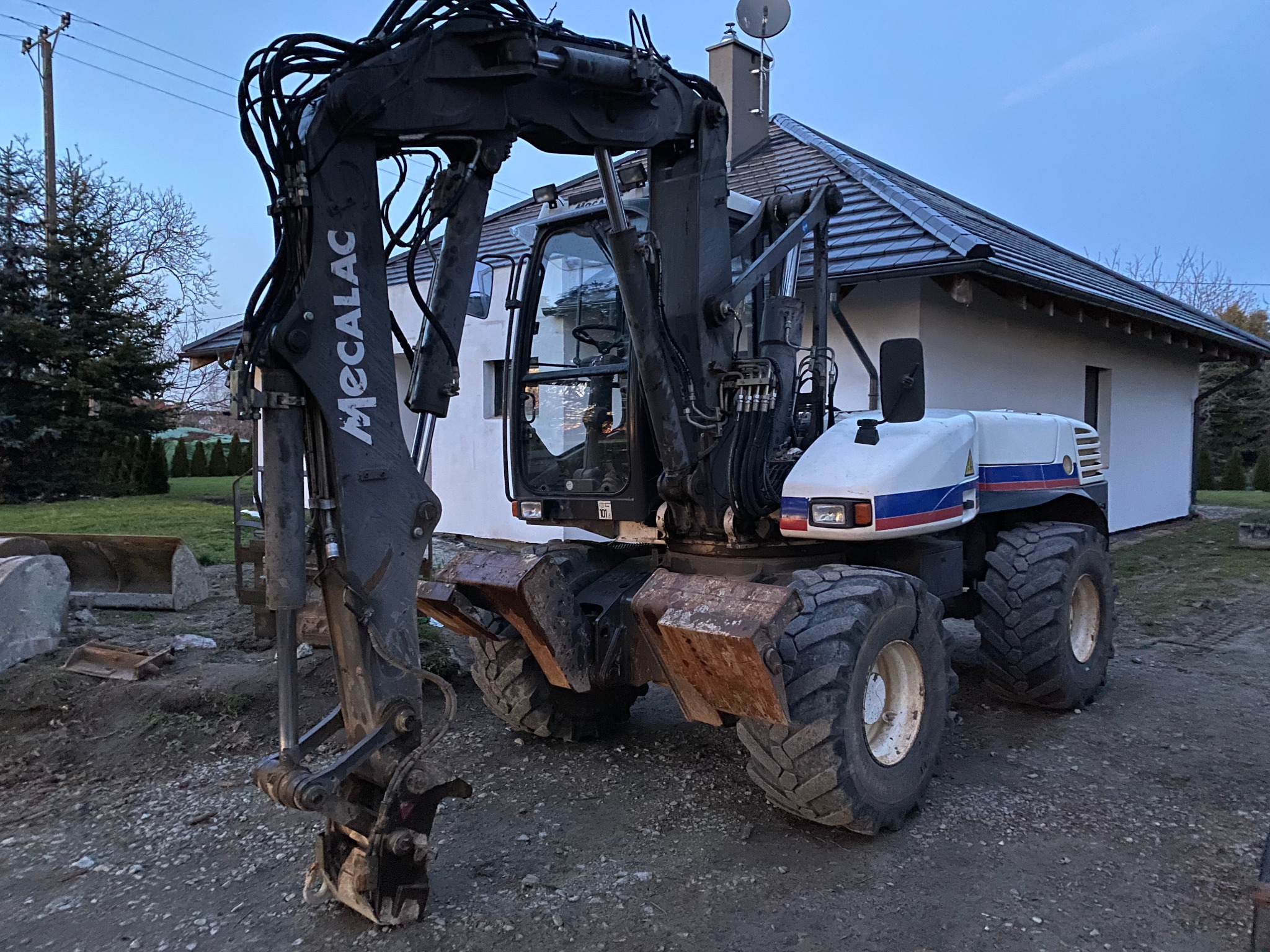 Ciężka maszyna Mecalac, prawdopodobnie koparka kołowa, stoi na gruncie w Oławie, z domem w tle. Widoczne przewody hydrauliczne i łyżka, sugerujące prace ziemne.