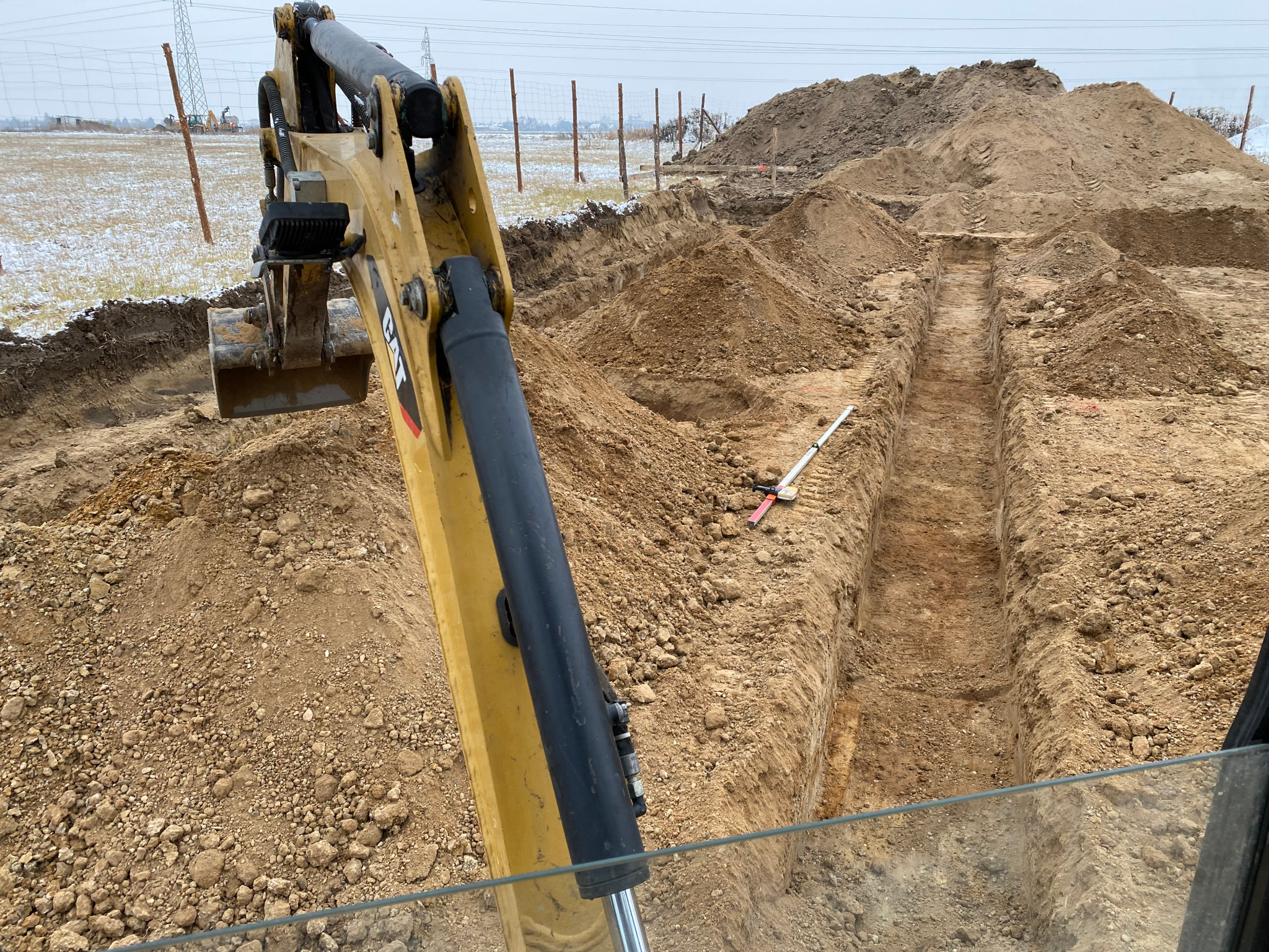 Żółta koparka z łyżką w trakcie prac ziemnych, widoczny wykop liniowy pod instalacje w ziemi, w tle słupy wysokiego napięcia i lekko ośnieżone pole.