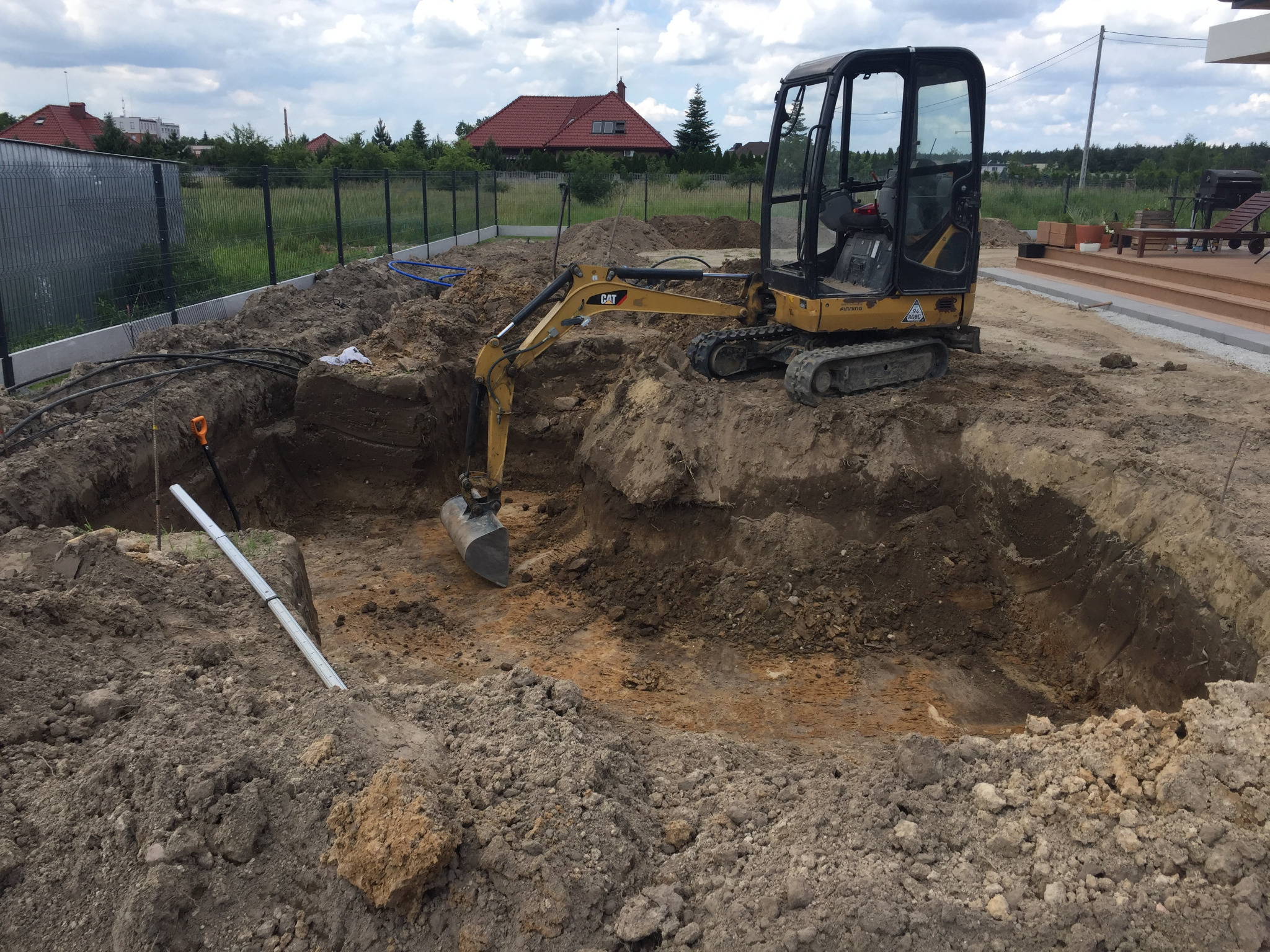 Żółta mini koparka CAT w wykopie, widoczne rury, teren przygotowany pod budowę, w tle dom z czerwonym dachem i ogrodzenie.