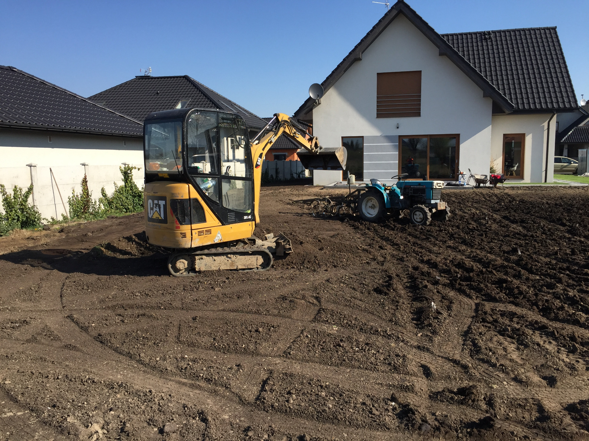 Żółta mini-koparka CAT 301.4C i mały niebieski traktor na wyrównanym, brązowym terenie budowy przed nowoczesnym domem z szarą elewacją i ciemnym dachem.