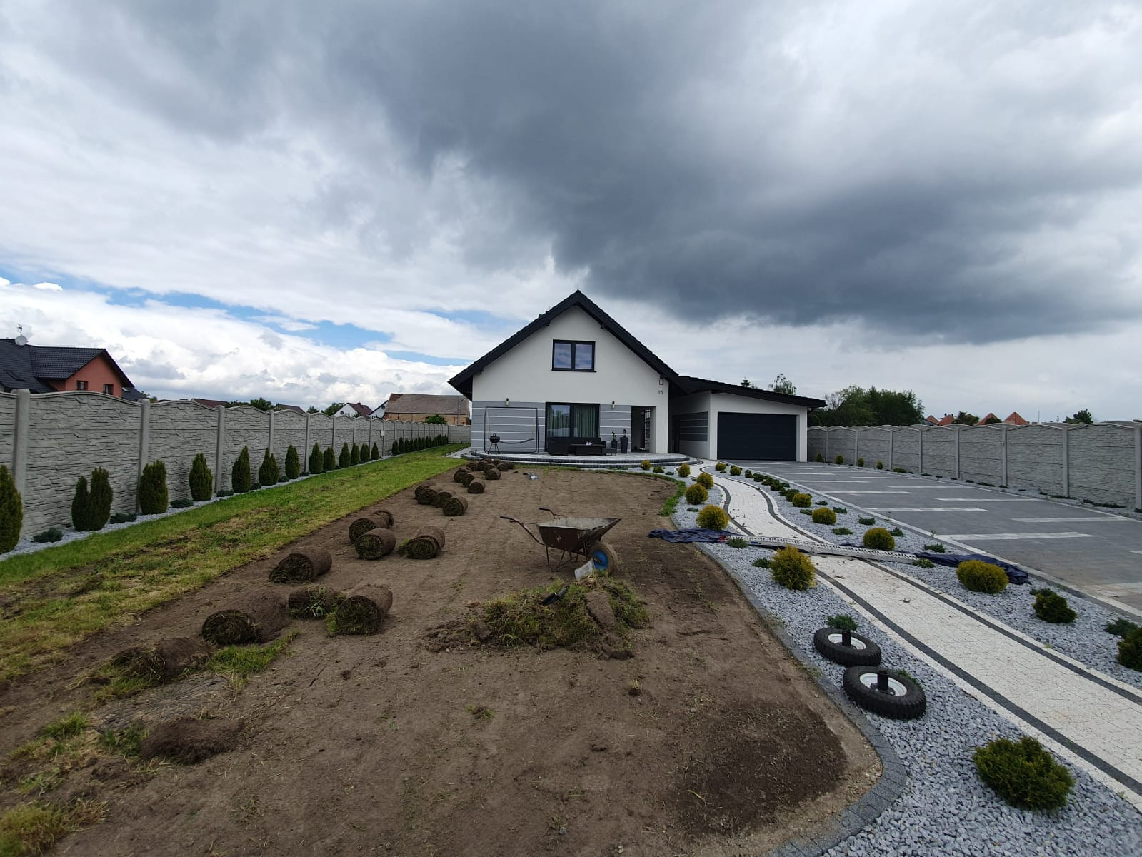 Przygotowanie terenu pod trawnik z rolki przed nowoczesnym domem, widoczne rozłożone rolki trawy, taczka z narzędziami, wykończony podjazd z kostki brukowej i ozdobne nasadzenia