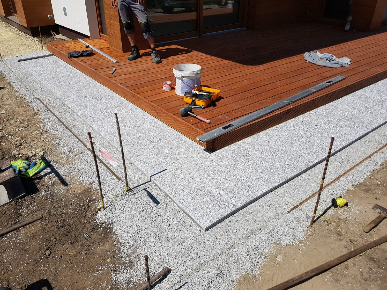 Jasnoszare płyty chodnikowe układane wokół drewnianego tarasu, widoczne narzędzia i materiały budowlane, częściowo wykończona powierzchnia z podsypką, poziomica na tarasie, pracownik w tle.