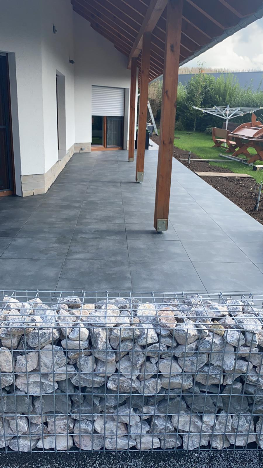 Zadaszony taras z szarych płyt, obudowany gabionem wypełnionym kamieniami, widok na zadbany ogród z suszarką na pranie i drewnianym stołem piknikowym.