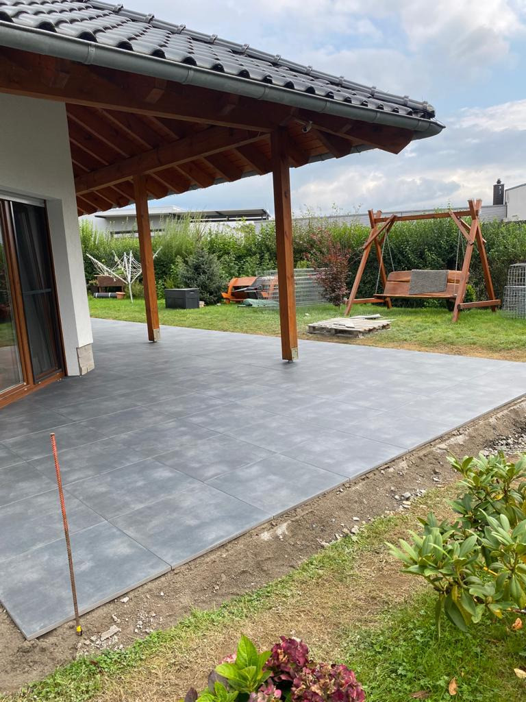 Nowoczesny taras z szarych płyt betonowych, częściowo ukończony, z drewnianą pergolą i huśtawką w tle, widok z poziomu gruntu.