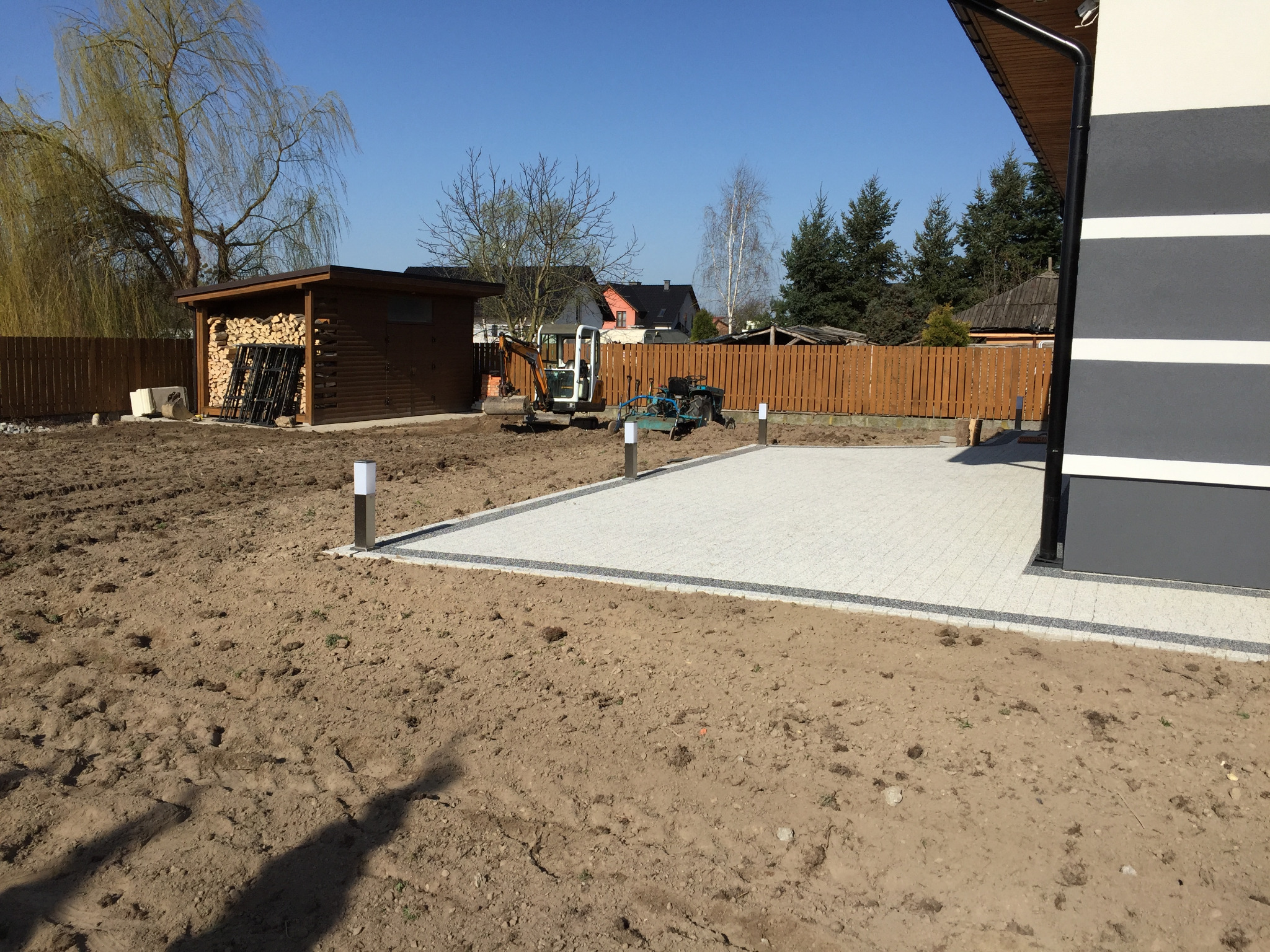 Ułożona kostka brukowa przy budynku z elementami szaro-białej elewacji, w tle przygotowany teren z mini koparką i traktorkiem oraz drewniana wiata na drewno.