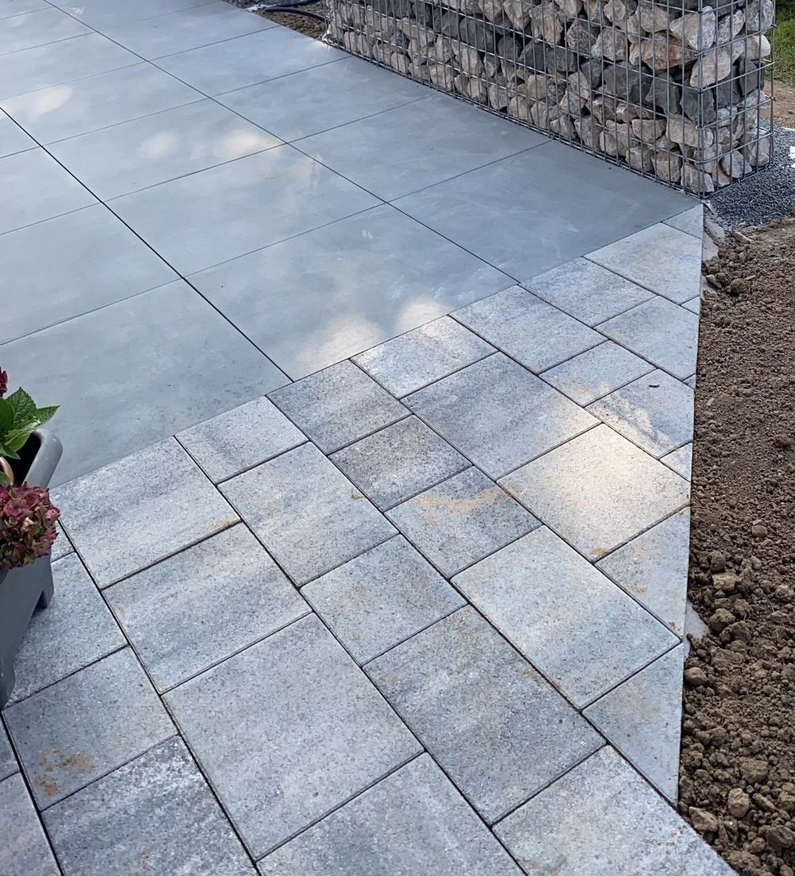 Dwa rodzaje szarej kostki brukowej na tarasie, po lewej stronie doniczka z kwiatami, w tle mur z gabionów wypełnionych kamieniami.