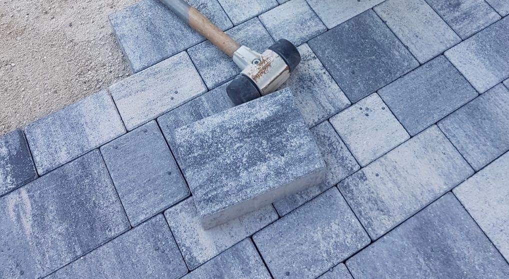 Układanie kostki brukowej z szaro-niebieskich elementów, widoczny gumowy młotek i podsypka piaskowa