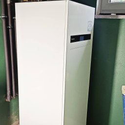 Bartosz Szewczyk Hydraulik Kraków - Biała jednostka wewnętrzna z niebieskim zbiornikiem wyrównawczym i rurami miedzianymi, zamontowana na tle zielonej ściany, widoczne okno i fragment dachu za oknem.