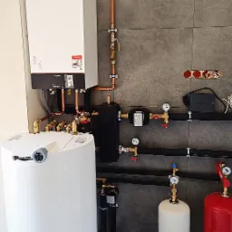 Biały kocioł grzewczy Viessmann z podłączeniami miedzianymi i zbiornikiem, na tle szarej ściany z płytek, widoczne manometry i zawory.