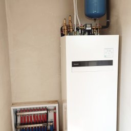 Bartosz Szewczyk Hydraulik Kraków - Instalacja pompy ciepła Panasonic Aquarea w surowym pomieszczeniu w Węgrzcach Wielkich, widoczny rozdzielacz ogrzewania podłogowego z rurami czerwonymi i niebieskimi oraz orurowanie do podłączenia.