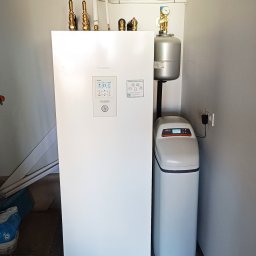 Bartosz Szewczyk Hydraulik Kraków - Biała pompa ciepła Panasonic Aquarea Smart Cloud z widocznymi przyłączami hydraulicznymi i zaworami, obok zmiękczacz wody Viessmann Aquahome 20, zainstalowane w pomieszczeniu technicznym z szarą...