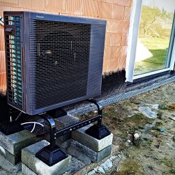 Bartosz Szewczyk Hydraulik Kraków - Zewnętrzna jednostka pompy ciepła Panasonic zamontowana na betonowych bloczkach przy ścianie budynku w trakcie budowy, widoczne ocieplenie fundamentów i drenaż.