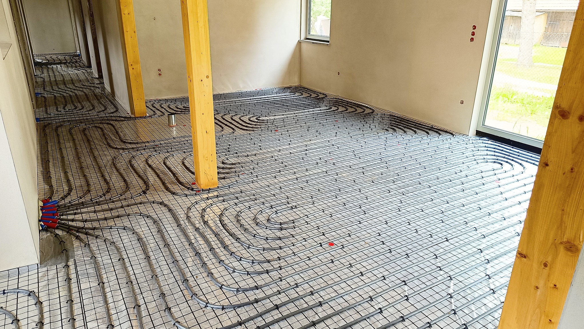 Rozłożone rury ogrzewania podłogowego na siatce w surowym stanie, widoczne drewniane podpory i okno z widokiem na zieleń.