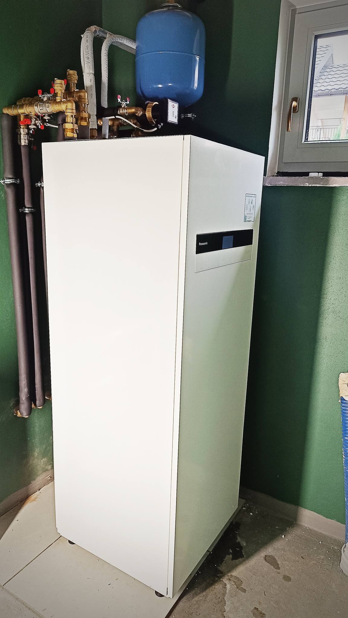 Biała jednostka wewnętrzna z niebieskim zbiornikiem wyrównawczym i rurami miedzianymi, zamontowana na tle zielonej ściany, widoczne okno i fragment dachu za oknem.