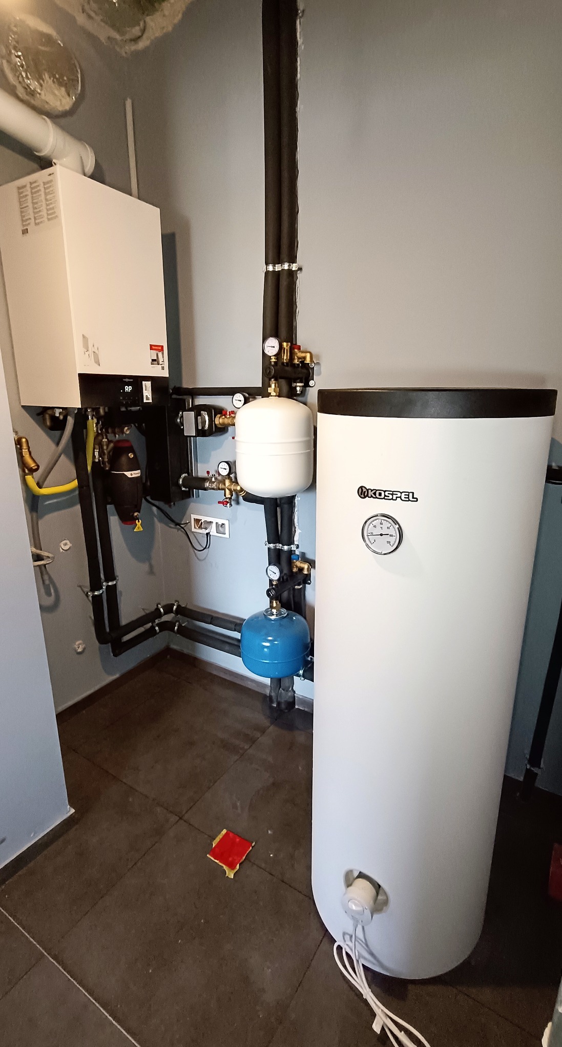 Biały zasobnik ciepłej wody użytkowej marki Kospel, z widocznym manometrem, podłączony do instalacji z czarnymi rurami izolowanymi, z dodatkowymi zbiornikami wyrównawczymi w kolorze białym...