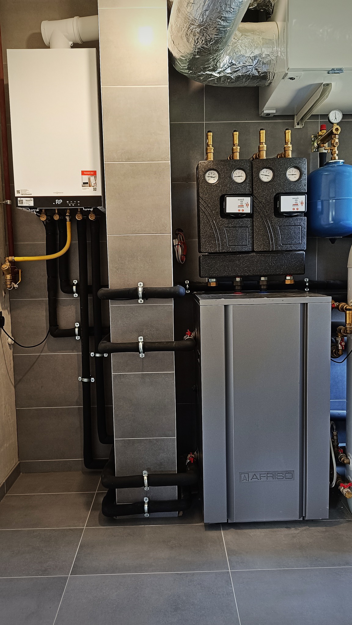 Nowoczesna kotłownia w Węgrzcach Wielkich z białym kotłem gazowym Viessmann, szarym zasobnikiem Afriso, czarnymi rurami instalacyjnymi i szarą glazurą ścienną i podłogową.