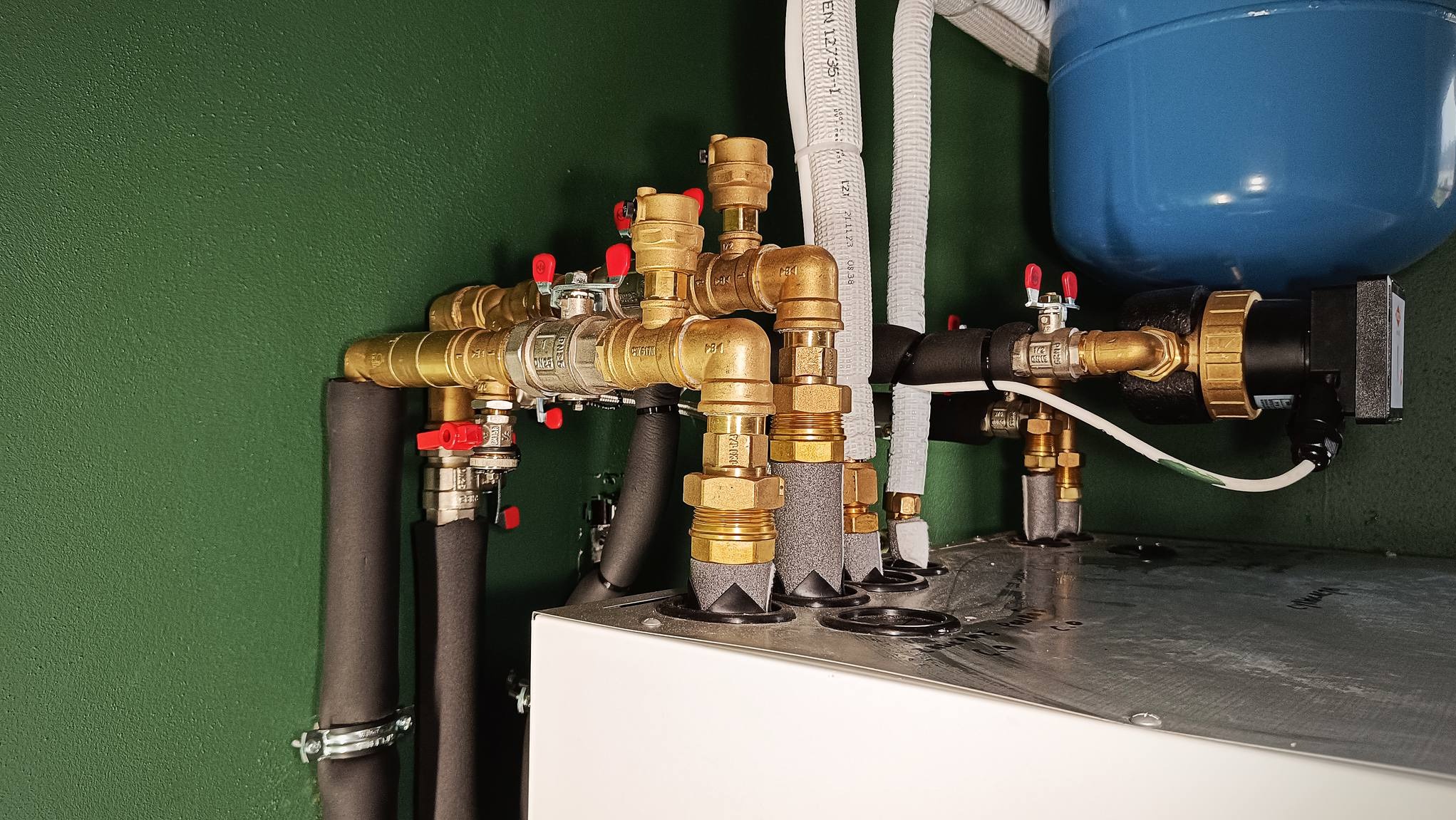 Kompleksowy węzeł instalacyjny z licznymi zaworami, rurami miedzianymi i izolacją, zamontowany na białej obudowie urządzenia na tle zielonej ściany.