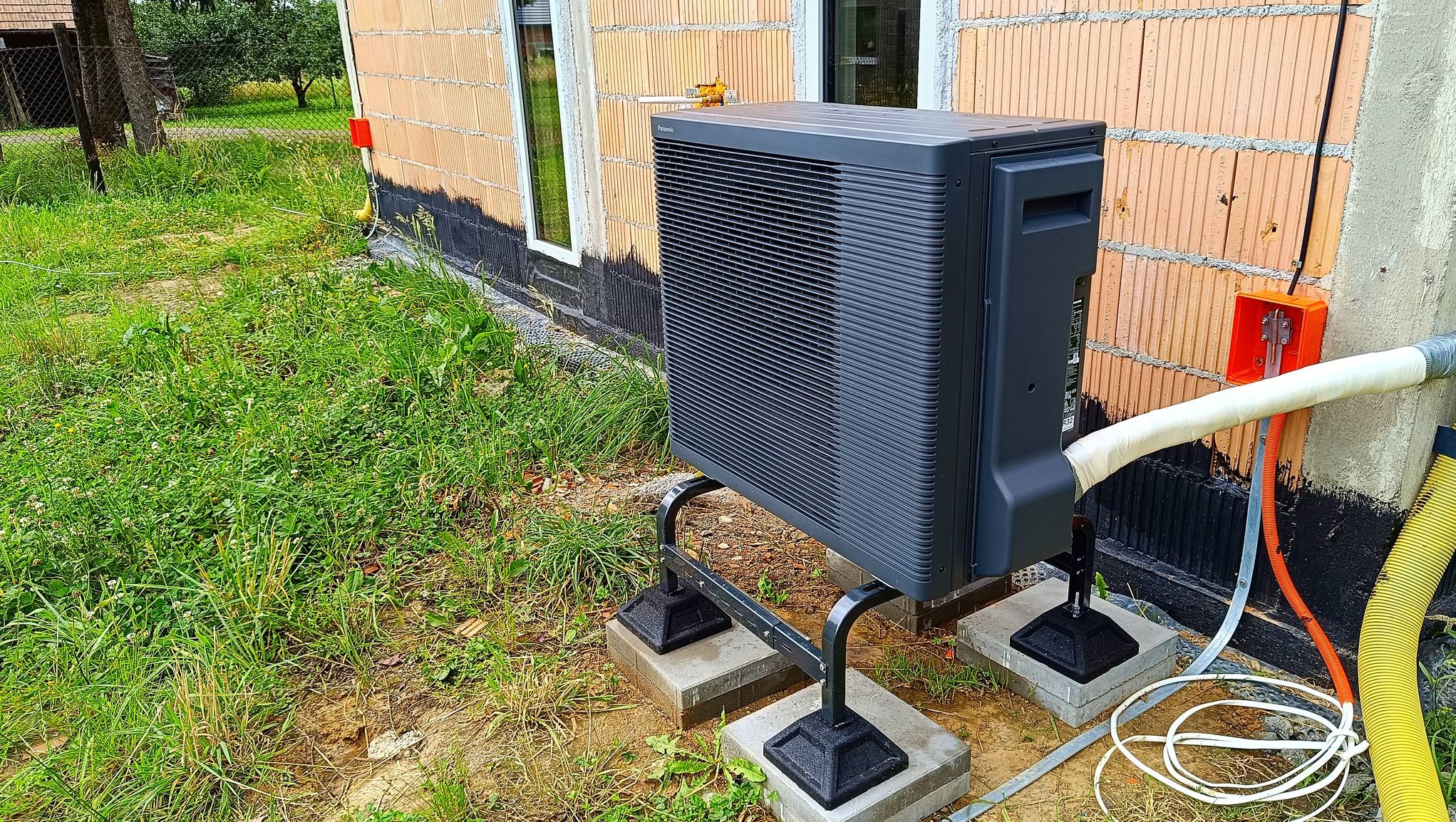 Zewnętrzna jednostka czarnej pompy ciepła Panasonic Aquarea, zamontowana na metalowym stelażu i betonowych podstawach, na tle surowej ściany budynku z cegły ceramicznej i bujnej trawy.