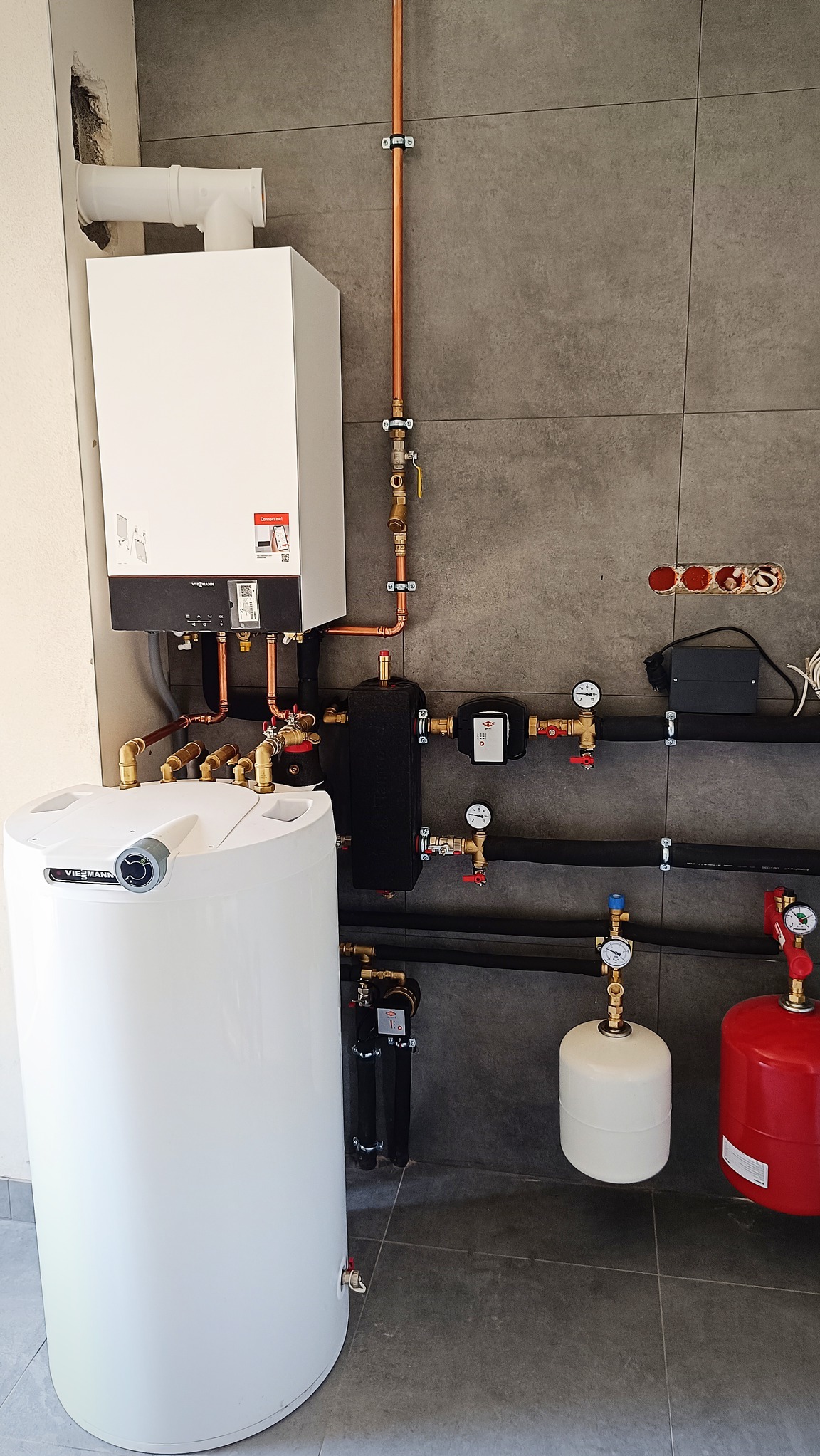 Biały kocioł grzewczy Viessmann z podłączeniami miedzianymi i zbiornikiem, na tle szarej ściany z płytek, widoczne manometry i zawory.