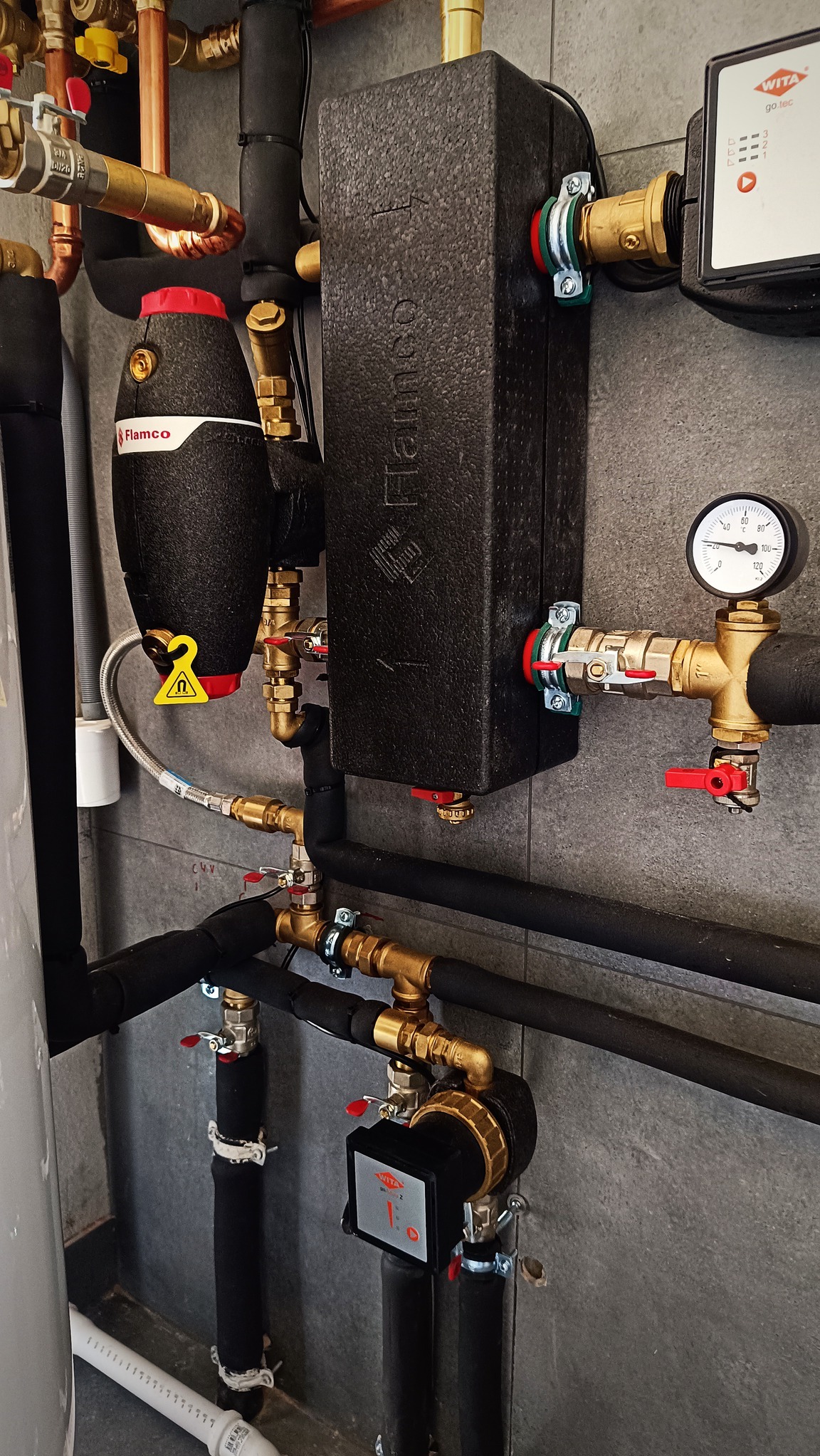 Czarny, izolowany zbiornik wyrównawczy Flamco z żółtą zawieszką ostrzegawczą i manometrem, zamontowany w systemie rur miedzianych i czarnych izolowanych, na tle szarej ściany z płytek.