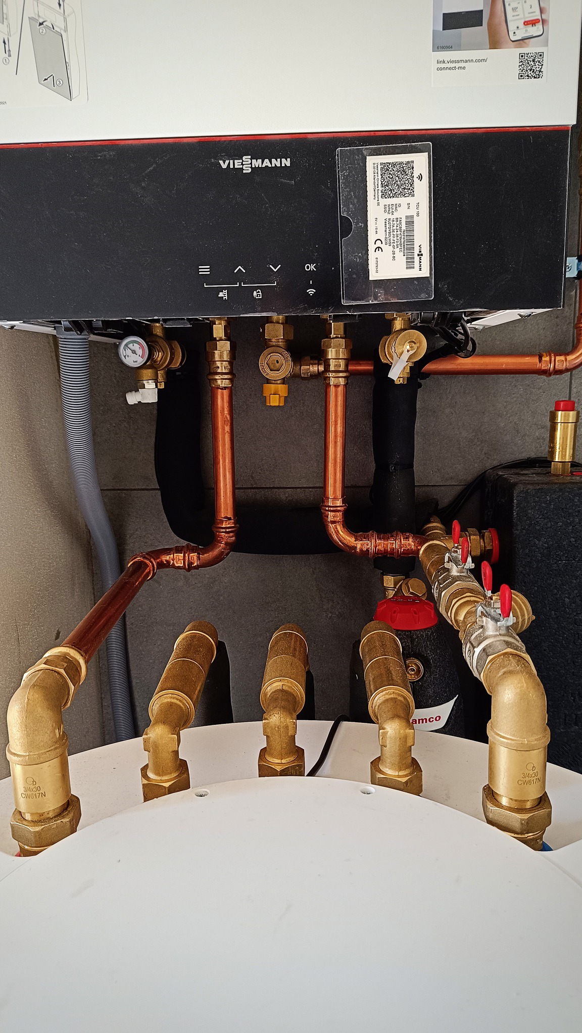 Czarny piec Viessmann z miedzianymi rurami i zaworami, widoczne przyłącza do instalacji grzewczej, z naklejką informacyjną i kodem QR.