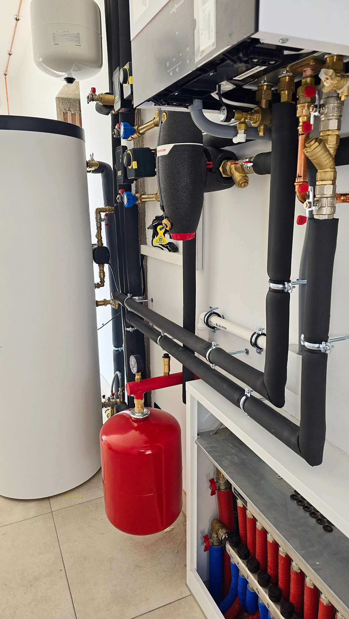 Nowoczesna kotłownia z białymi ścianami, widoczny piec Viessmann, zasobnik ciepłej wody, czerwony zbiornik wyrównawczy, czarne rury izolowane termicznie i rozdzielacz podłogówki...