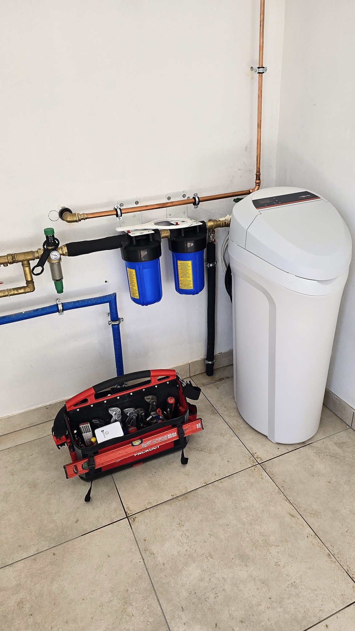 Biały zmiękczacz wody podłączony do instalacji z miedzianych rur i niebieskich filtrów, obok otwarta czerwona skrzynka narzędziowa Milwaukee Packout z widocznymi narzędziami, na tle białej ściany...