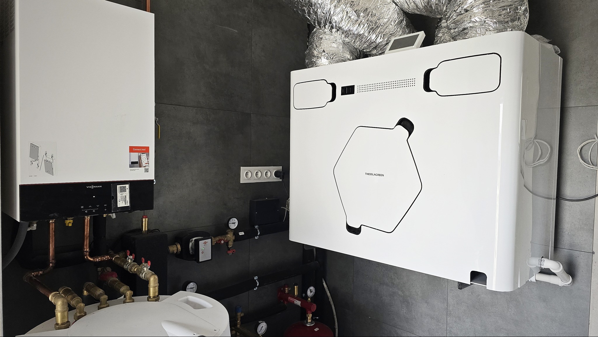 Biała pompa ciepła Thesslagreen z widocznymi przewodami wentylacyjnymi i kotłem Viessmann na szarej ścianie z płytek.
