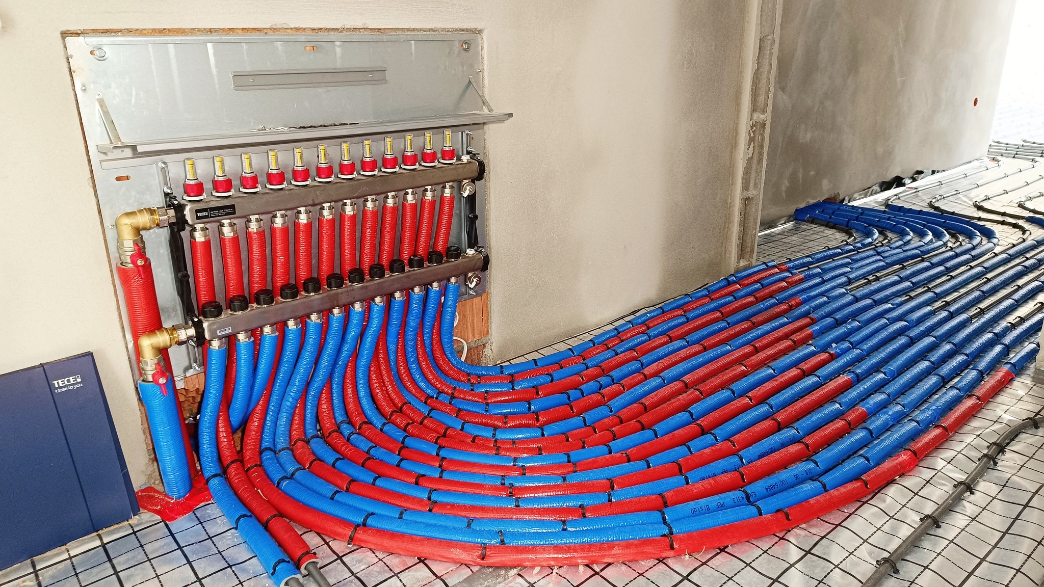 Rozdzielacz instalacji ogrzewania podłogowego z czerwono-niebieskimi rurami przymocowanymi do siatki, widoczne elementy metalowe i fragment ściany w surowym stanie.