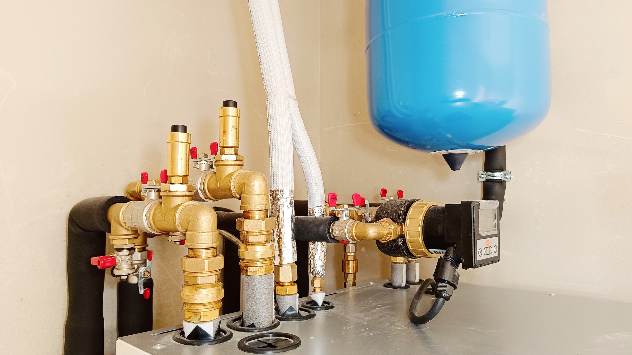 Kompleksowy system rur i zaworów z niebieskim zbiornikiem ciśnieniowym, izolowane rury, widoczne elementy instalacji grzewczej, zamontowane na metalowej konstrukcji przy jasnej ścianie.