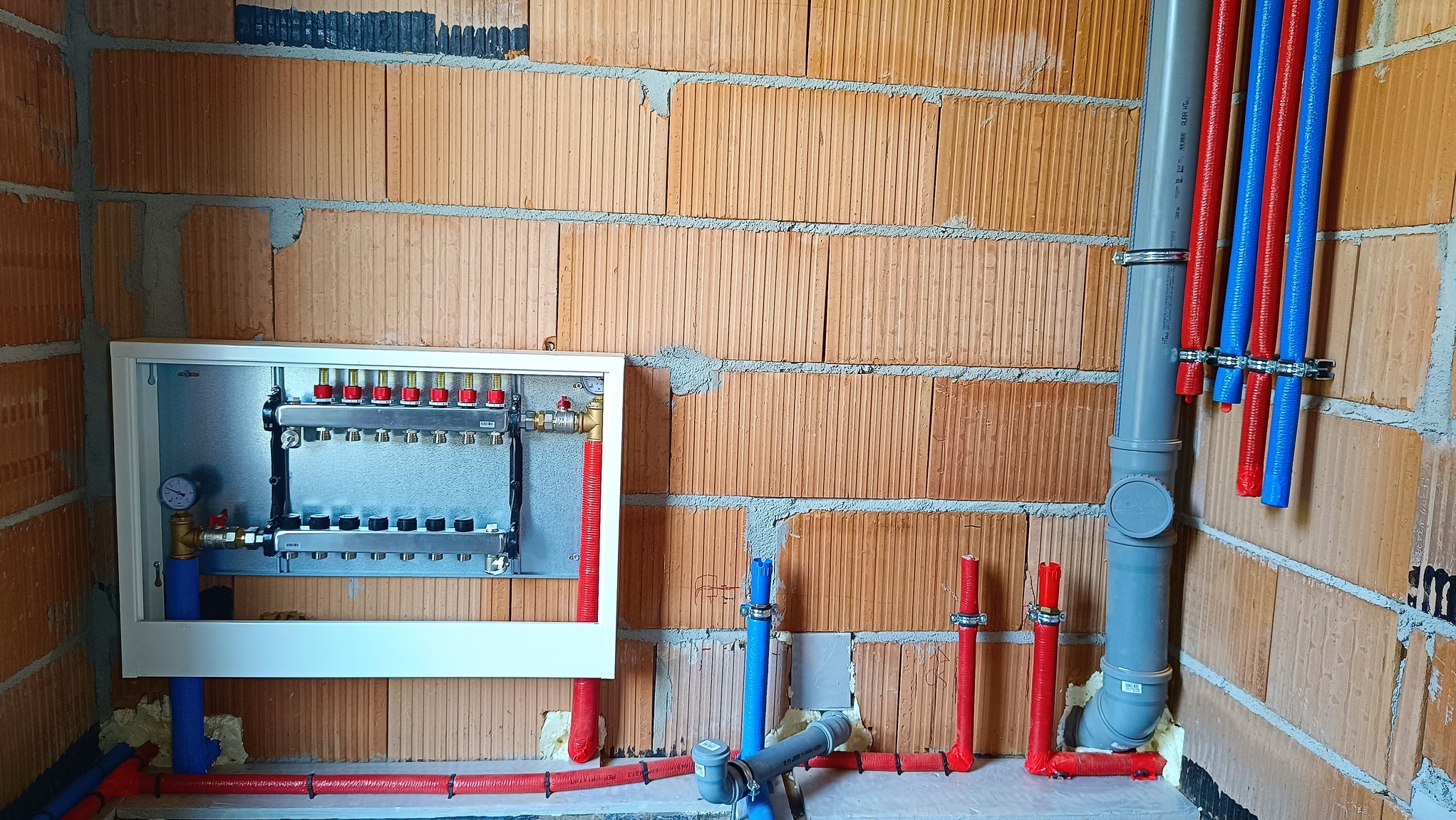 Rozdzielacz ogrzewania podłogowego w obudowie, z podłączonymi rurami czerwonymi i niebieskimi, zamontowany na ścianie z cegły kratówki.