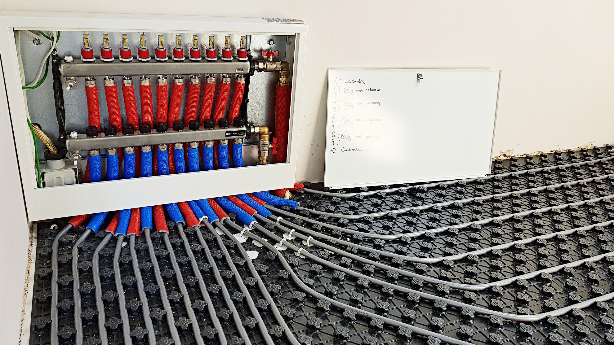 Rozdzielacz ogrzewania podłogowego z rurami w kolorze czerwonym i niebieskim, ułożonymi na czarnej macie izolacyjnej, obok biała tablica z listą pomieszczeń.