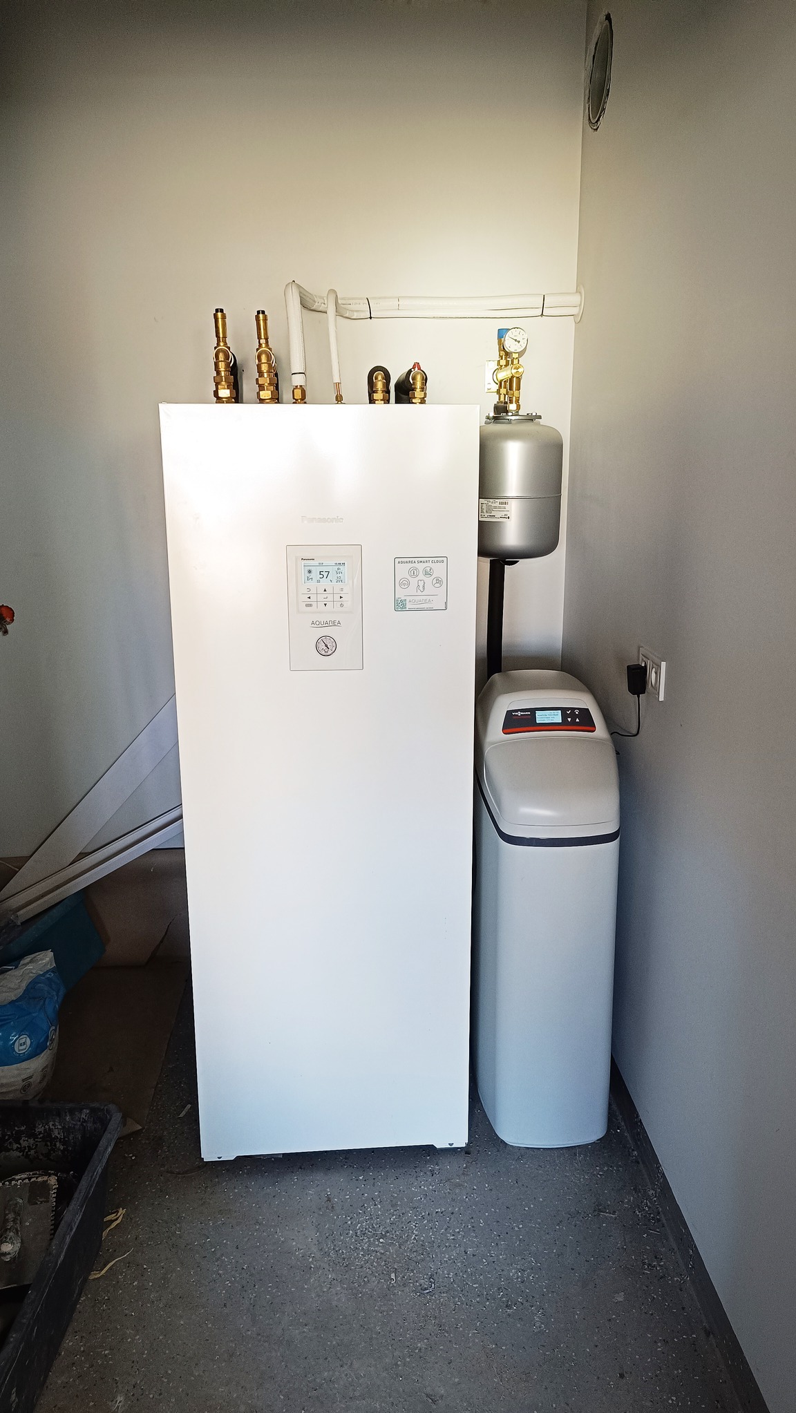 Biała pompa ciepła Panasonic Aquarea Smart Cloud z widocznymi przyłączami hydraulicznymi i zaworami, obok zmiękczacz wody Viessmann Aquahome 20, zainstalowane w pomieszczeniu technicznym z szarą...
