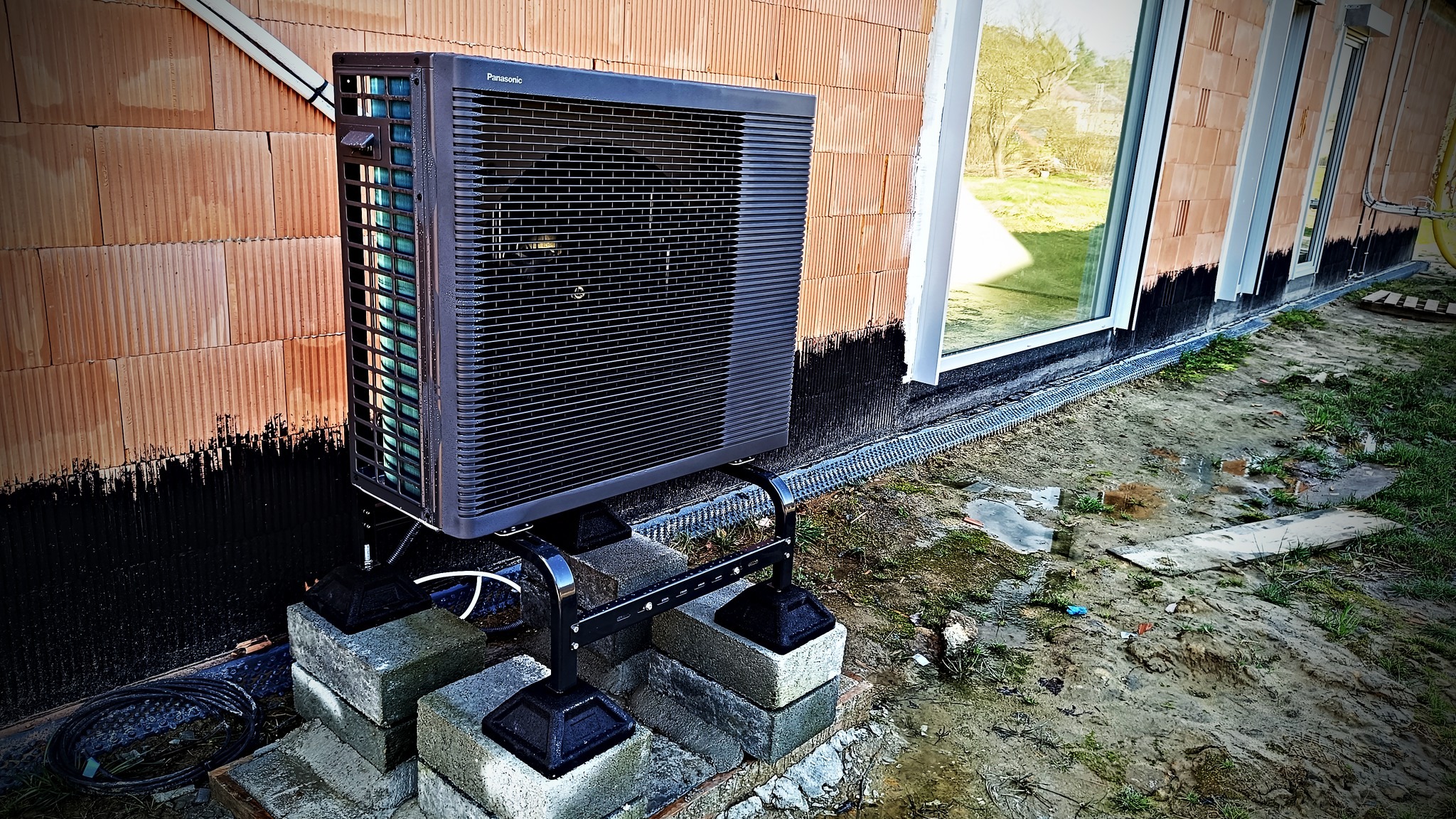 Zewnętrzna jednostka pompy ciepła Panasonic zamontowana na betonowych bloczkach przy ścianie budynku w trakcie budowy, widoczne ocieplenie fundamentów i drenaż.